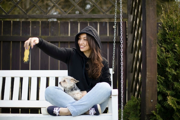 Attractive brunette woman with long hair dressed in black hoodie sitting on white swing bench with yorkshire dog and makes selfies with her smartphone