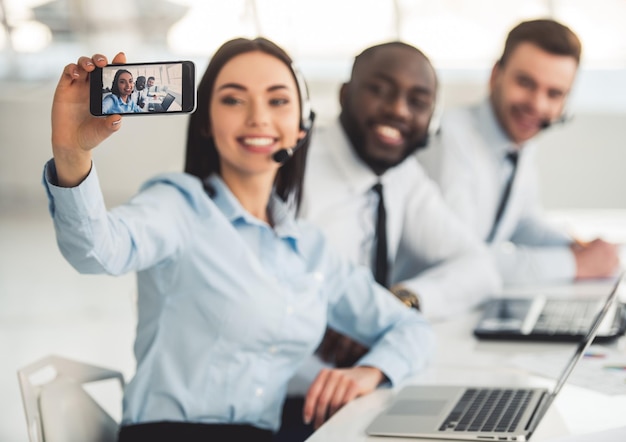 Attractive business people in suits and headsets are smiling while working with laptops in office Girl is doing selfie