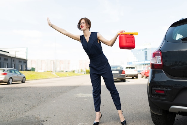 Привлекательная бизнес-леди держит красную канистру с бензиновым топливом на стоянке и протягивает руку с просьбой о помощи