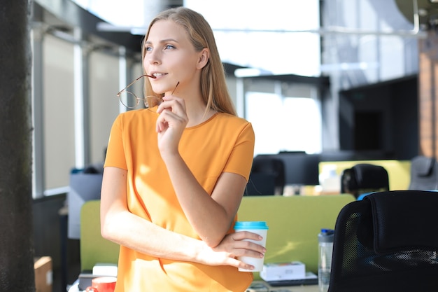 Attractive business woman is looking in the window and holding holding her glasses while standing in the office.