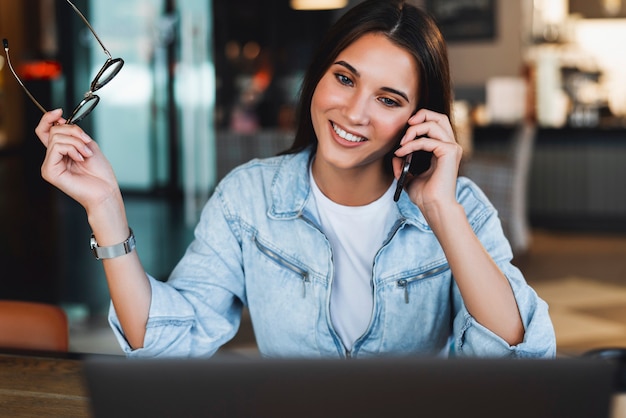 Attractive business woman sits at table in front of laptop, talks on mobile phone, negotiates on phone.