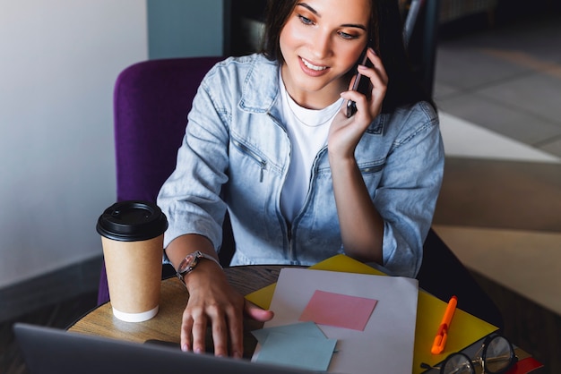 Attractive business woman sits at table in front of laptop and talks on mobile phone, negotiates on the phone.