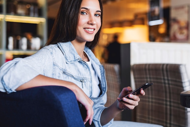 Attractive business woman sits at table and holds mobile phone in her hand. Portrait of beautiful brunette