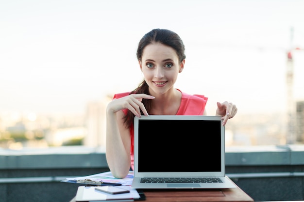 Привлекательная бизнес-леди в розовой блузке сидит на крыше и работает с ноутбуком, показывает пустой ноутбук