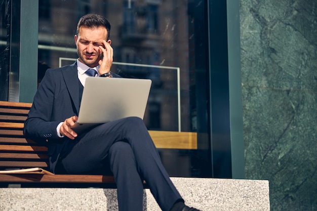 Attractive businessman sitting on bench alone outdoors with laptop on knees and touching head