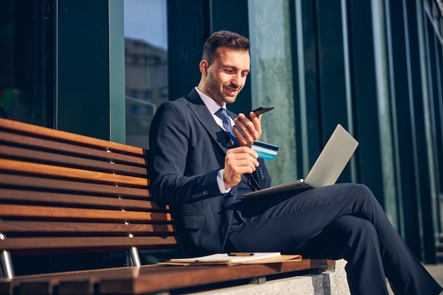 Attractive businessman sitting outside on bench while making call and holding credit card in hands