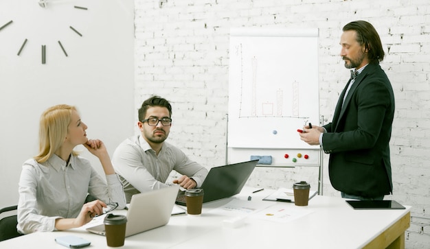 Attractive businessman standing near a white board presents to the team his business idea. Colleagues carefully listen to speaker sitting at the office table.