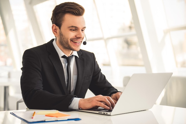 L'uomo d'affari attraente in vestito e auricolare sta sorridendo mentre lavora con il computer portatile in ufficio