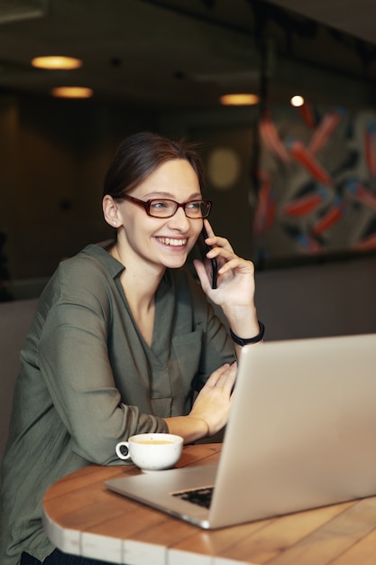 Attractive businesswoman in black jacket and stylish glasses sitting in cafe, talking on the phone and looking at laptop.