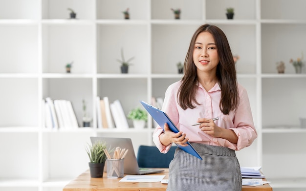 Attractive businesswoman holding file folder standing at office smiling to camera