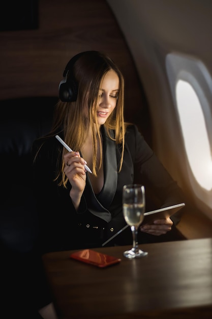 Attractive businesswoman using digital tablet in private plane.
lovely caucasian young woman in stylish black dress listening music
with wireless headphones and working during flight.