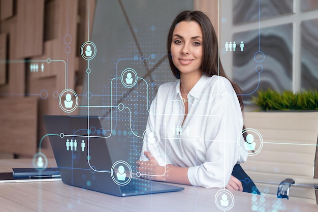 Attractive businesswoman in white shirt at workplace working with laptop to hire new employees for international business consulting HR social media hologram icons over office background