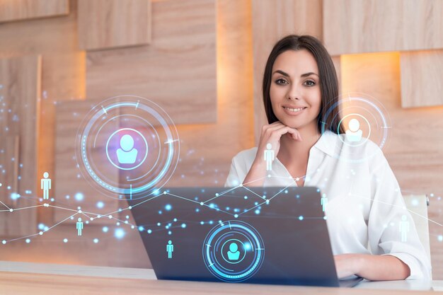 Attractive businesswoman in white shirt at workplace working\
with laptop to hire new employees for international business\
consulting hr social media hologram icons over office\
background