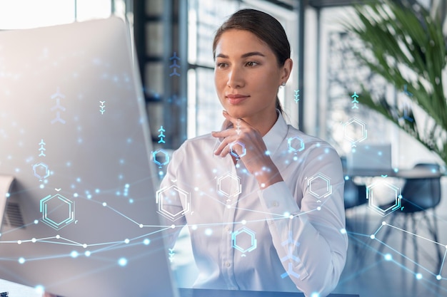 Attractive businesswoman in white shirt at workplace working with laptop to optimize development by implying new technologies in business process Hi tech hologram over office background