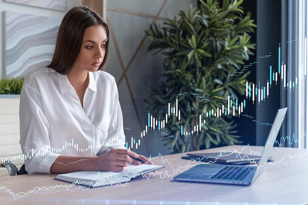 Attractive businesswoman in white shirt at workplace working\
with laptop to optimize trading strategy at corporate finance fund\
forex chart hologram over office background