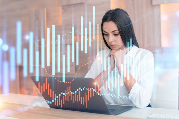 Attractive businesswoman in white shirt at workplace working with laptop to optimize trading strategy at corporate finance fund Forex chart hologram over office background