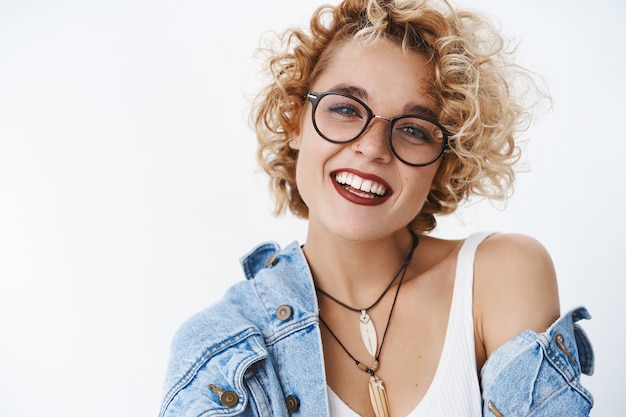 Attractive carefree and lovely hipster woman with blond hair in glasses and lipctick smiling charming at front and gazing happily enjoying romantic date on valentines day over white wall