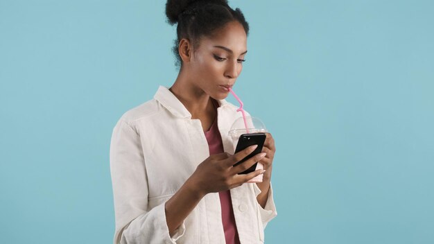 스마트폰을 열심히 사용하고 화려한 배경 위에 밀크셰이크를 마시는 매력적인 캐주얼 아프리카계 미국인 소녀