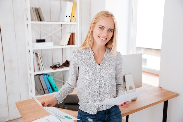 Attractive casual businesswoman holding documents and sitting on the office desk