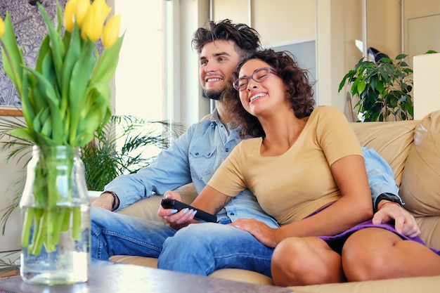 The attractive casual couple sits on a couch and switching tv channels with remote control.