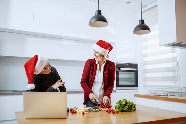 魅力的な白人金髪女性が野菜を刻んで、健康的なクリスマスディナーを準備します。彼女の隣に立って、料理の仕方をアドバイスする母親。どちらも頭にサンタ帽子をかぶっています。