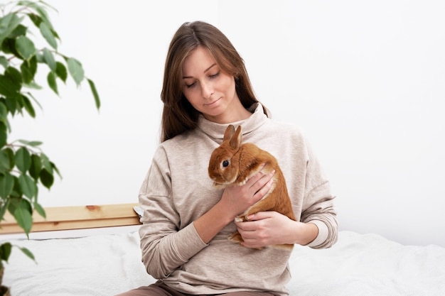 Attractive caucasian brunette womangirl with long hair\
holdingplaying with cute little bunny rabbit rodentsitting on bed\
at home on white backgroundpet care animal loveeaster concept