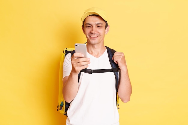 Attractive Caucasian delivery man in yellow cap and tshirt with thermal backpack holding cell phone looking at display chatting while has free time isolated on yellow background