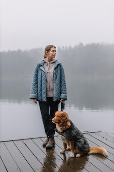 Attraente ragazza caucasica in giacca blu che resta sul molo con nova scotia duck tolling retriever marrone. donna e cane che viaggiano insieme. nebbia sopra l'acqua. alberi sullo sfondo. gli animali amano il concetto.