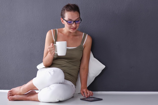 Attractive caucasian girl sitting on floor with cup and tablet near wall