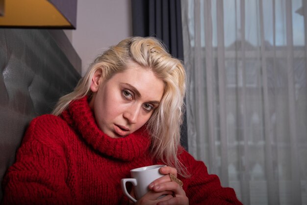 Attractive caucasian girl wears a heavy red sweater, it is winter and she is indoor and drinks a hot drink from the cup. Eastern european girl in a winter indoor mood concept