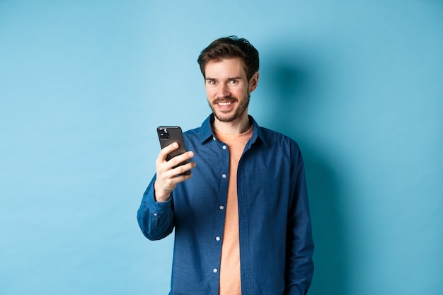 Attractive caucasian man using mobile phone, looking happy and smiling at camera, standing on blue background.