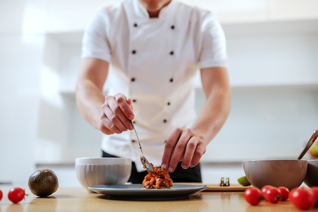 Attractive Caucasian serious chef in uniform putting prepared meal on plate.