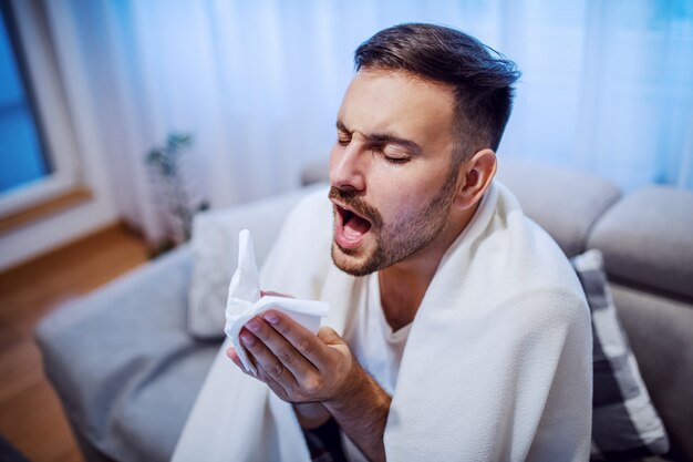 Attractive caucasian unshaven man sitting on sofa in living room covered with blanket, holding handkerchief and sneezing.