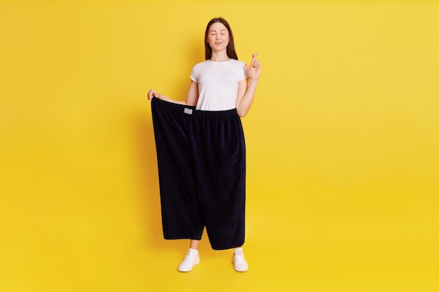 Attractive Caucasian young female wearing old huge pair of pants, weight loss, stands with close eyes, keeps fingers crossed, make wish to not get fat again, isolated over yellow wall.