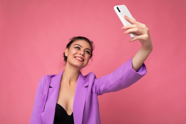 魅力的な魅力的な若い幸せな女性は、壁の背景に隔離されたスタイリッシュな服を着て自分撮りを取っている携帯電話を保持し、使用しています。