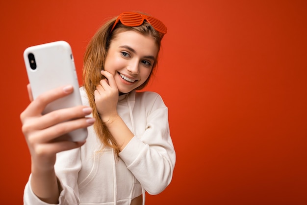 Attractive charming young smiling happy woman holding and using mobile phone taking selfie wearing