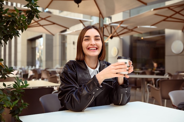 Attractive cheerful brunette european woman with cardboard\
coffee cup smiles toothy smile looking at camera, resting in summer\
terrace of outside cafeteria restaurant, enjoying autumn cool\
weather.