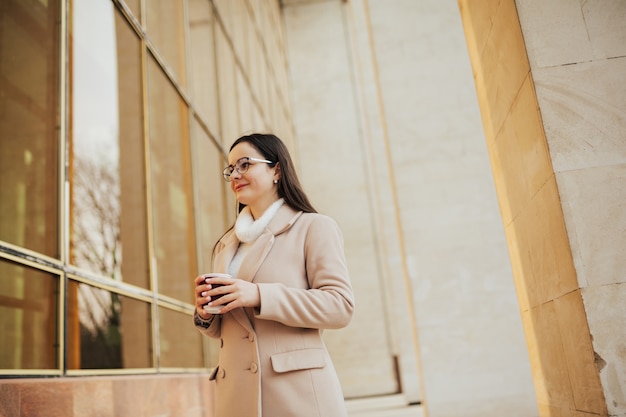 Attractive cheerful girl in beige coat and eyeglasses with coffee to go walking outdoor near building.