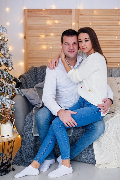 Attractive cheerful man and woman sitting near christmas tree and hugging each other. New Year's eve.