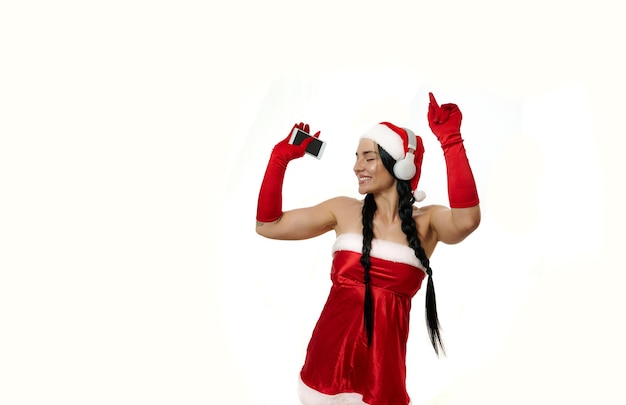 Attractive cheerful playful woman in Santa Claus carnival costume wearing wireless headphones, holding smartphone and dancing against white wall background with copy space for Christmas advertisement