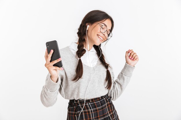 Attraente studentessa allegra che indossa unifrom in piedi isolato su un muro bianco, ascoltando musica con gli auricolari, tenendo il telefono cellulare, ballando