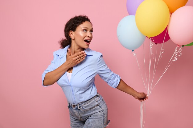 Attractive cheerful surprised woman of mixed race ethnicity looks at multicolored bright air balloons in her hands, isolated over pink background with copy space for text and advertising