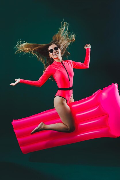 Attractive cheerful woman in a pink swimsuit on an isolated background she has an inflatable mattress between her legs Vacation concept Slender girl in sunglasses on an air mattress