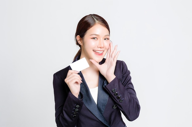 Attractive cheerful young Asian businesswoman wearing formal suit and smiling while standing and holding credit card on isolated white background in studio