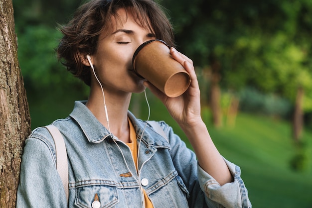 Привлекательная жизнерадостная молодая девушка в повседневной одежде проводит время на открытом воздухе в парке, слушает музыку в наушниках и держит чашку кофе на вынос