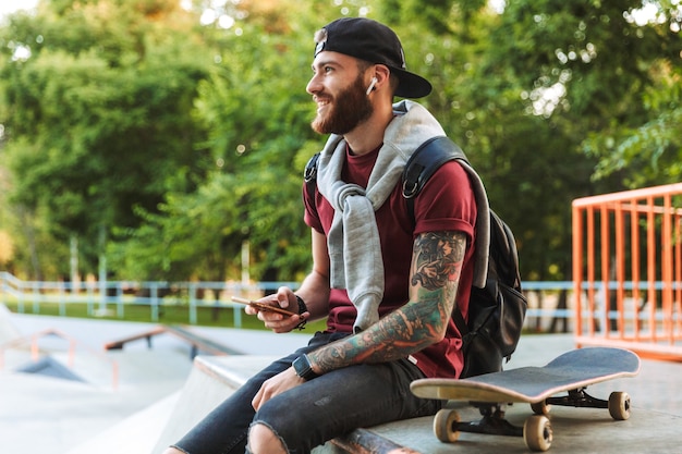 Attractive cheerful young man sitting at the skate park ramp with a skateboard, using mobile phone