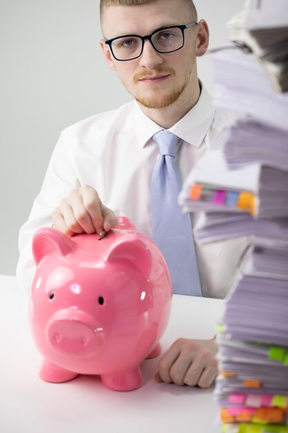 Attractive concentrated man inserting coin in piggybank in office, finance