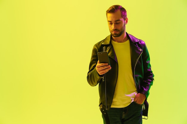 Attractive confident young man wearing leather jacket standing isolated over yellow wall, using mobile phone