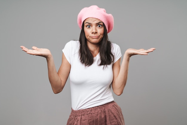 Attractive confused young brunette woman wearing beret standing isolated over gray wall, presenting copy space, shrugging shoulders