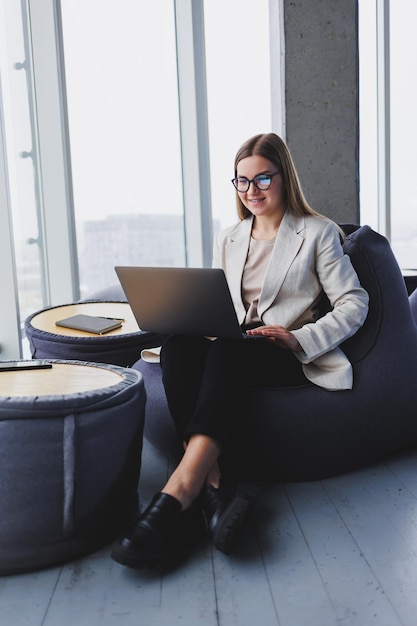 Attractive content blonde woman in casual wear and glasses concentrating on screen and typing on laptop while sitting on soft pouf in lounge area